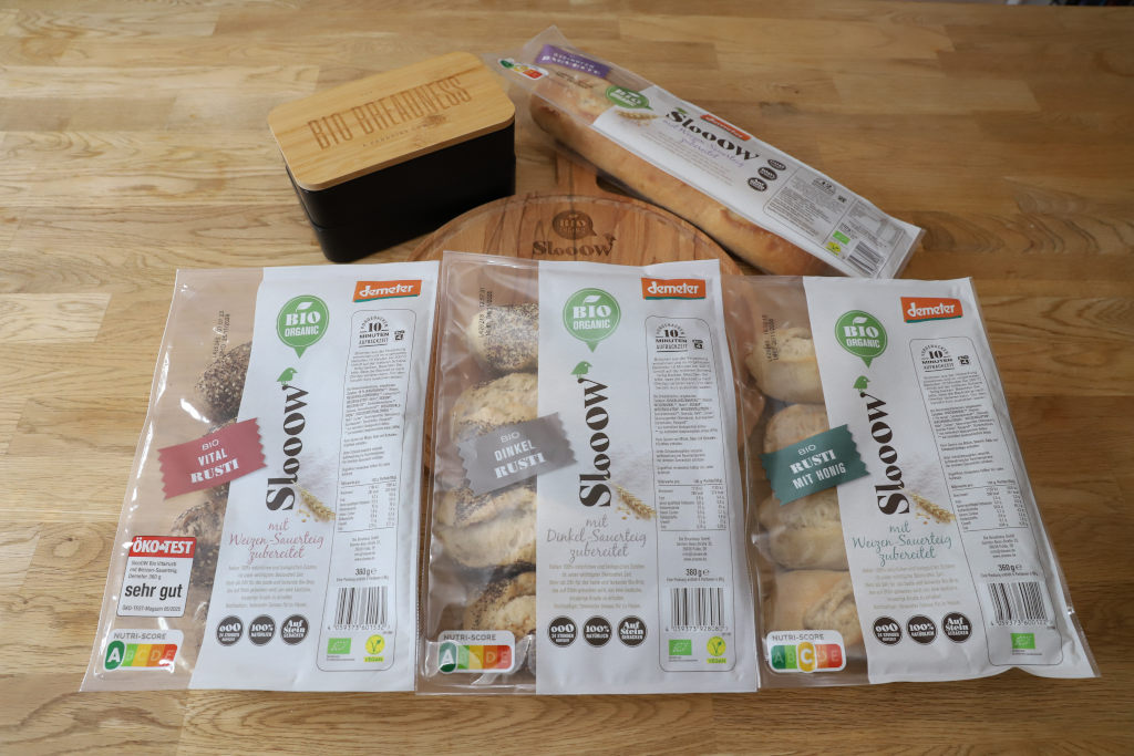 über bio“ drei Pakete mit je einer Packung Vital Rusti, Dinkel Rusti, Rusti mit Honig, Steinofen Baguette der Marke SlooOW und dazu das passende Holzschneidebrett sowie Brotdose aus dem Hause Bio Breadness
